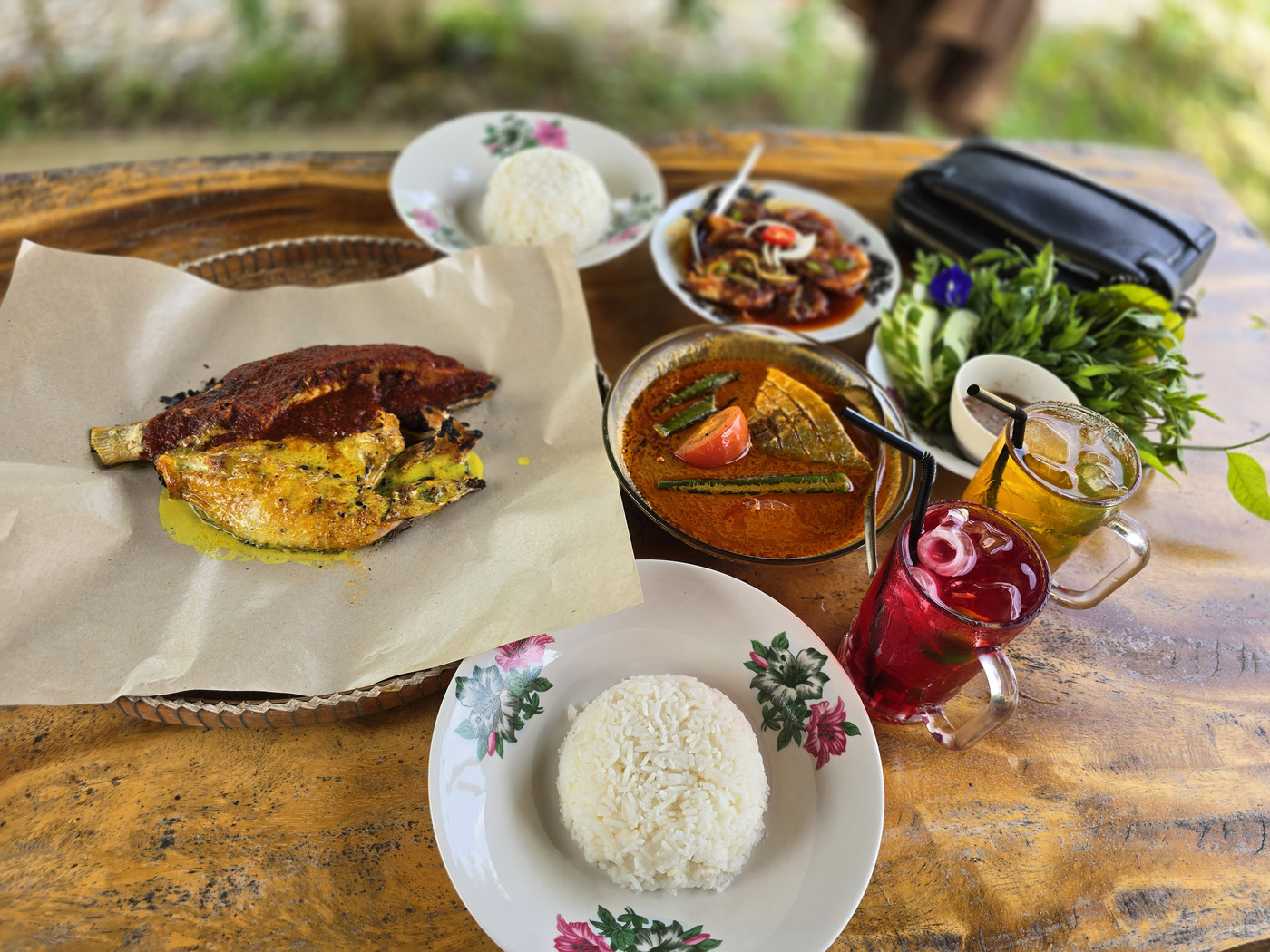 Grilled fish, curry, rice, raw vegetables and two iced drinks on a wooden table.