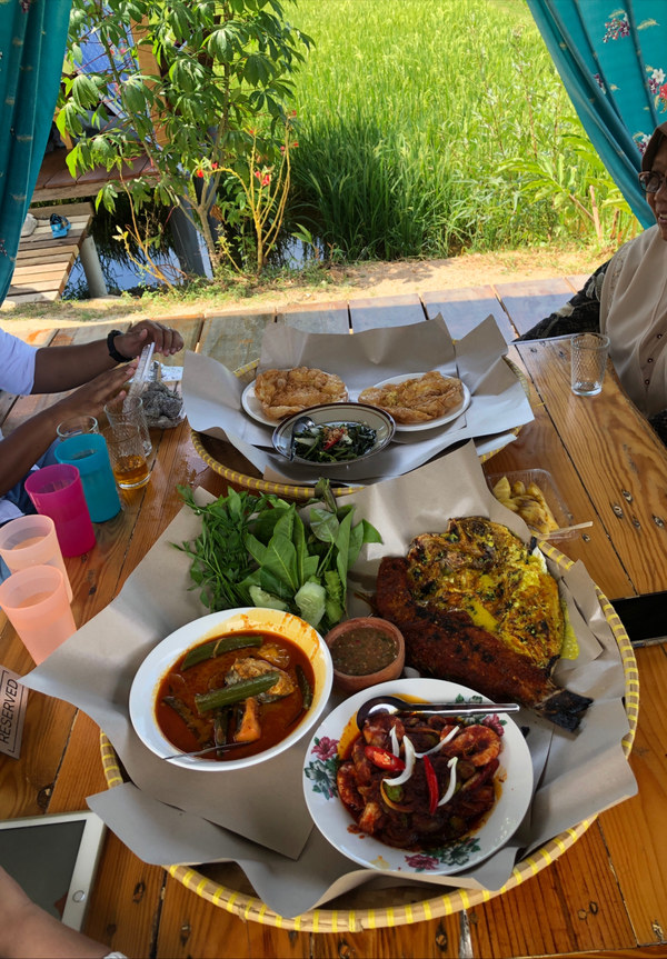 A large shared platter of grilled fish, curry, vegetables and side dishes beside a paddy field.