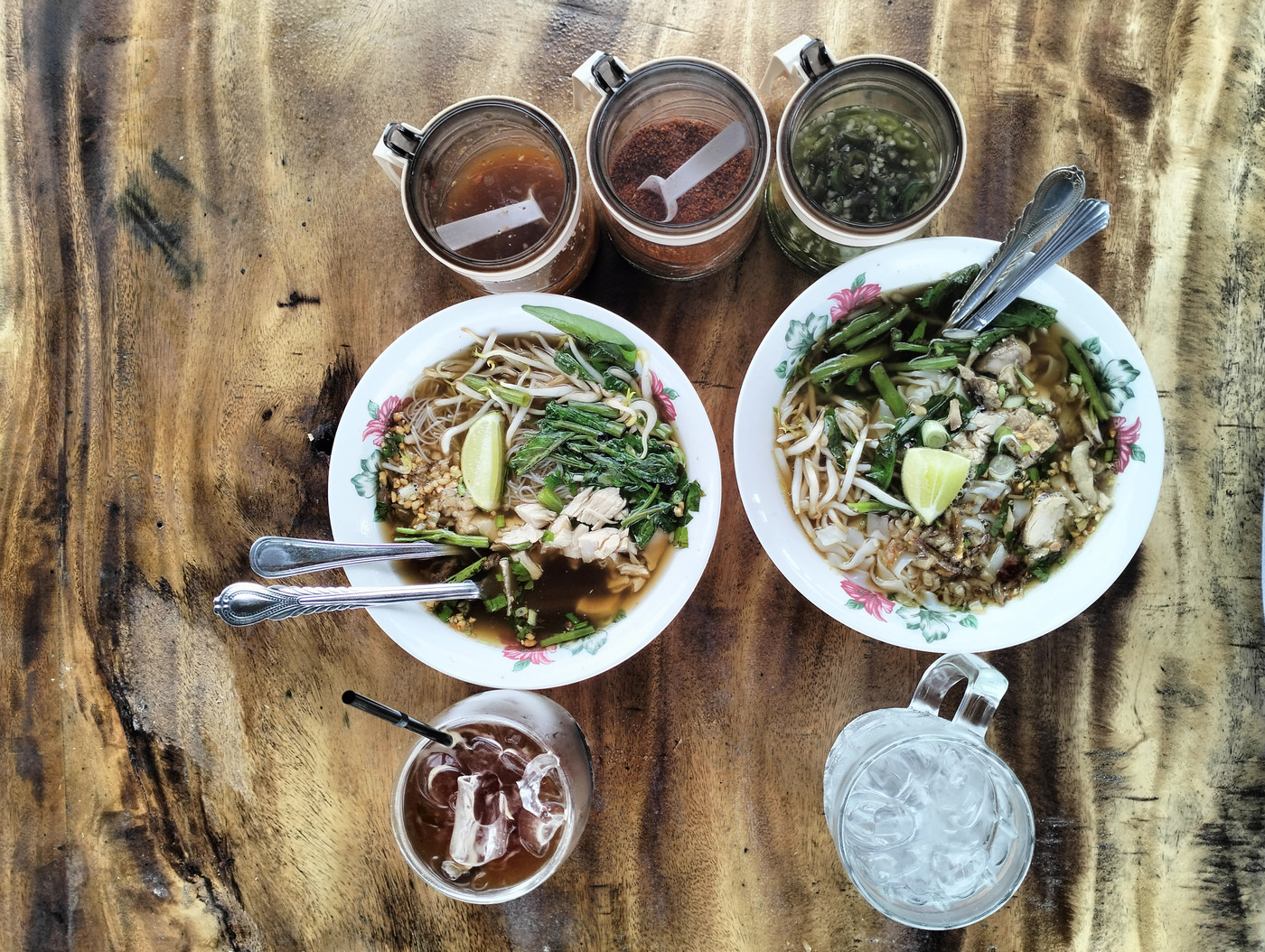 Two bowls of chicken noodle soup with condiments and iced drinks.