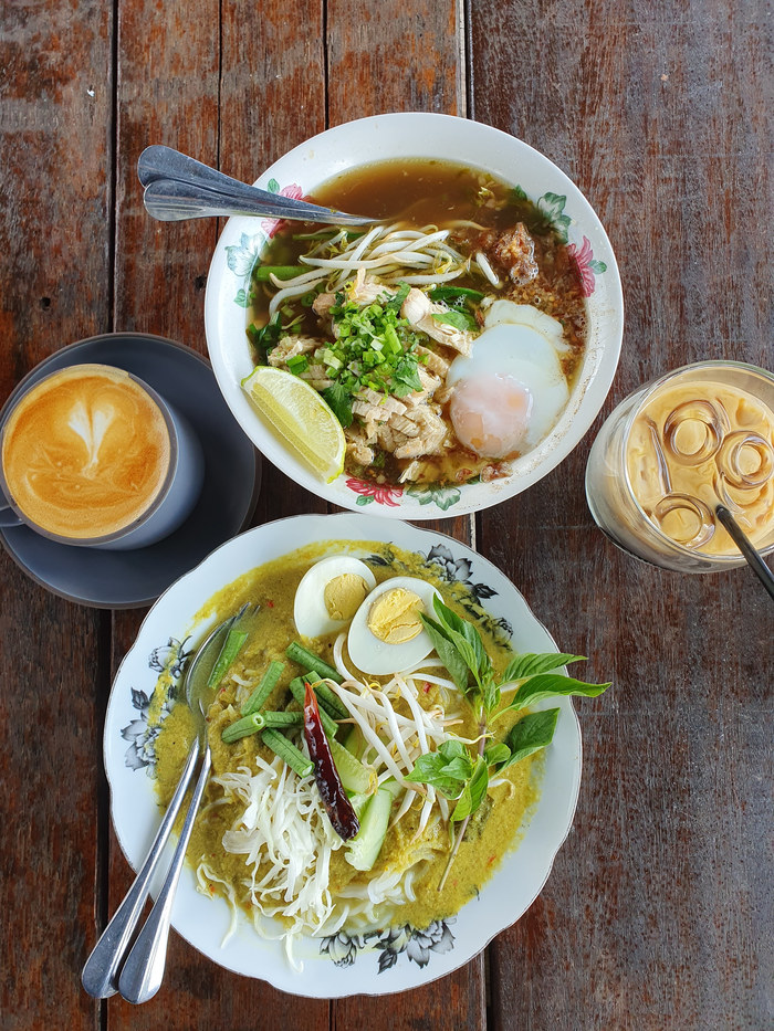 Two noodle dishes with coffee and an iced drink on a wooden table.