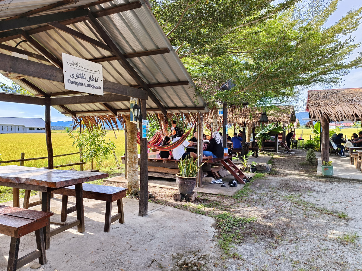 Open-air dining shelters beside a paddy field, with wooden tables and diners.